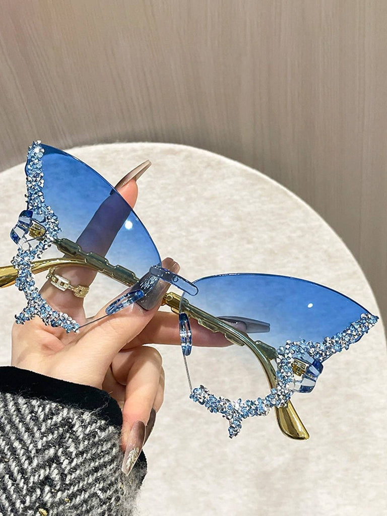 Blaue, schmetterlingsförmige Sonnenbrille mit goldenen Akzenten, die von einer Hand gehalten wird, vor einem neutralen Hintergrund