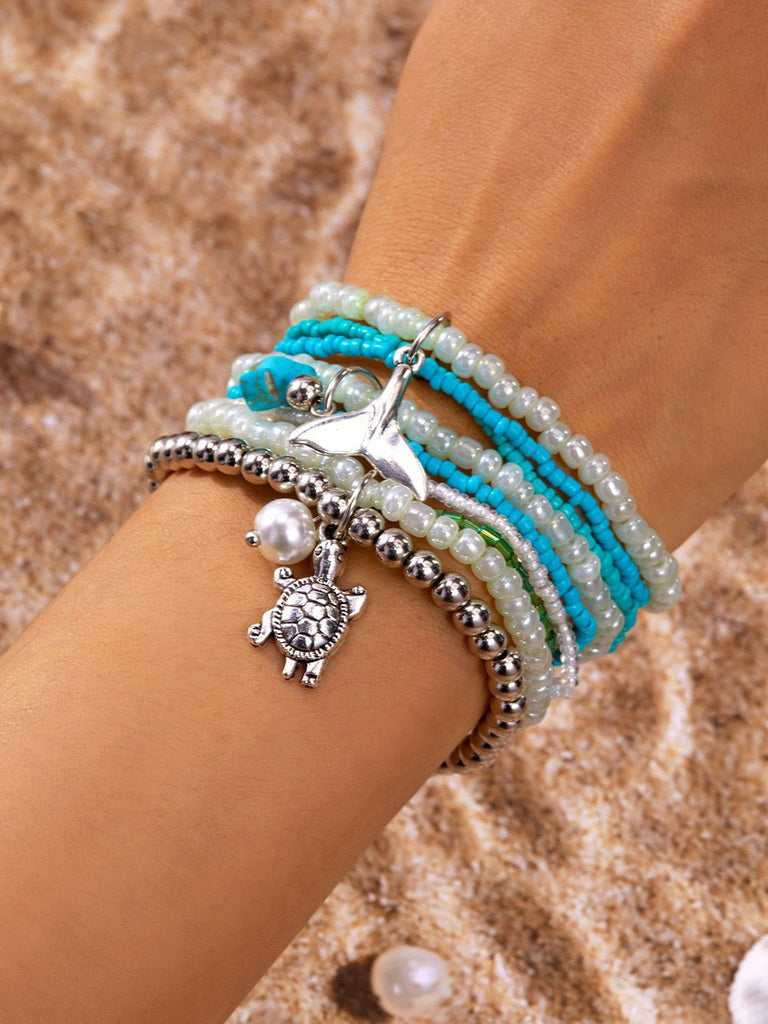 Stapel von Perlenarmbändern mit Schildkrötenanhänger am Handgelenk vor sandigem Hintergrund