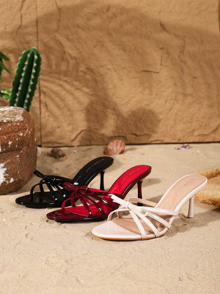 Schwarze, weiße und bordeauxrote Riemchen-Stiletto-Mules mit Knotenverschluss liegen zusammen auf einem sandigen Untergrund.