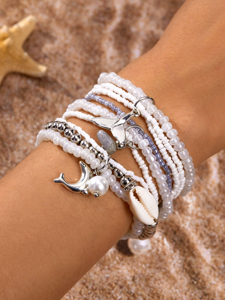 Mehrere Perlenarmbänder mit Muschel- und Metallanhängern an einem Handgelenk vor sandigem Hintergrund